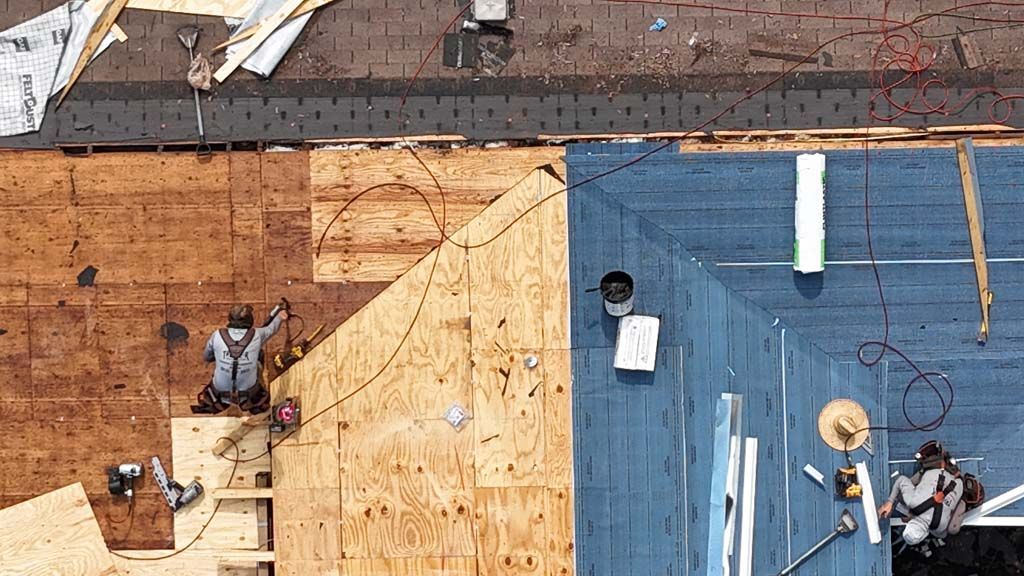 Roofers working on a roof, some areas with new plywood, some with blue covering, overhead shot.