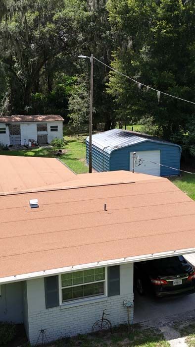 Brown-roofed house with a car in the carport, a blue shed, and a utility pole in a yard with trees.