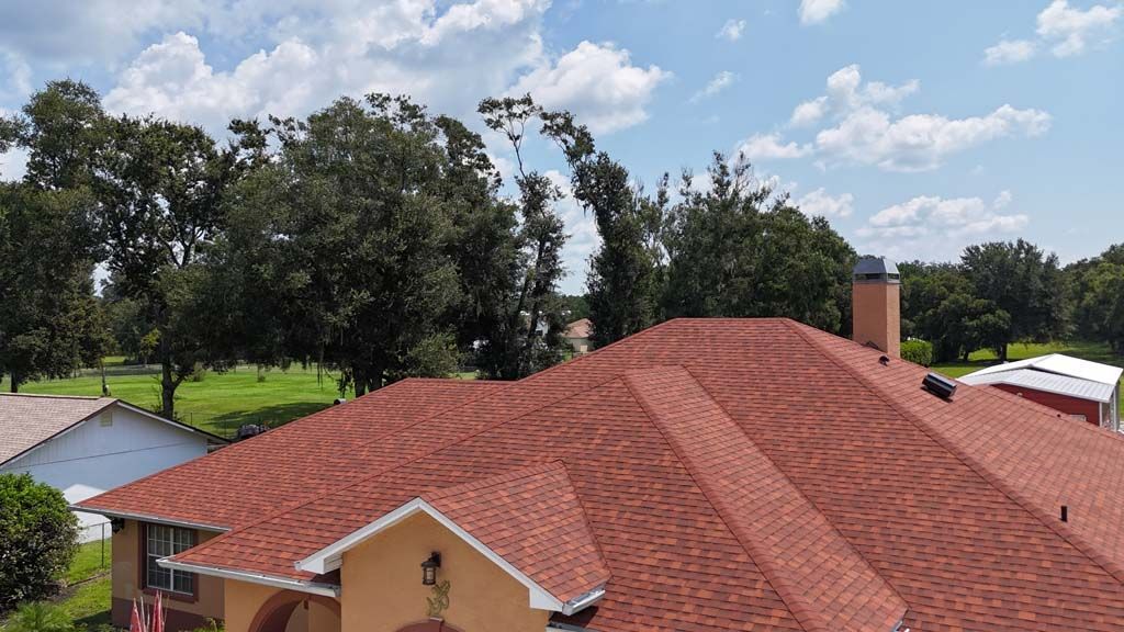Red shingle roof of a house, with chimney and surrounding trees under a partly cloudy sky.