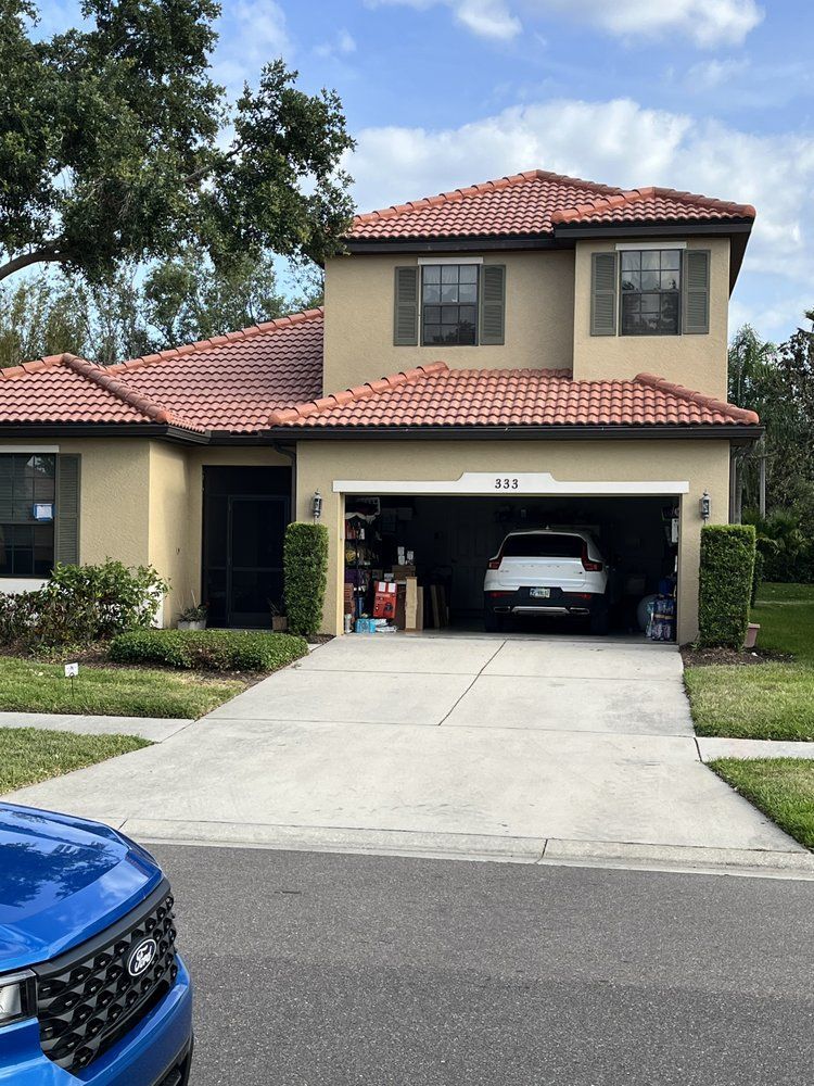 Two-story beige house with a red tile roof, car in open garage, blue car in foreground.