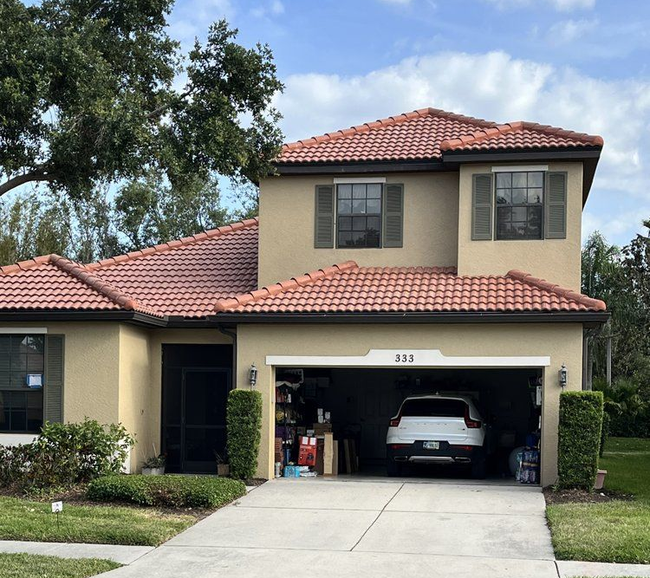 Tan two-story house with red tile roof, attached garage with a car inside, and green shutters.
