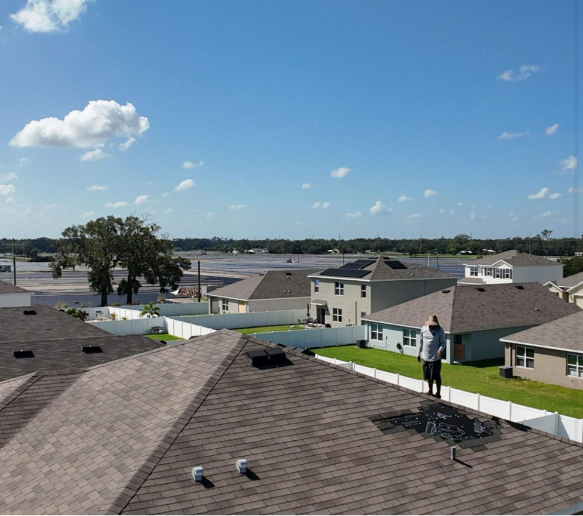 Man on a roof, checking for damage; residential neighborhood with blue sky and fluffy clouds.