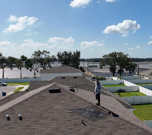 Person on a roof inspecting shingles on a sunny day with a view of houses and trees.