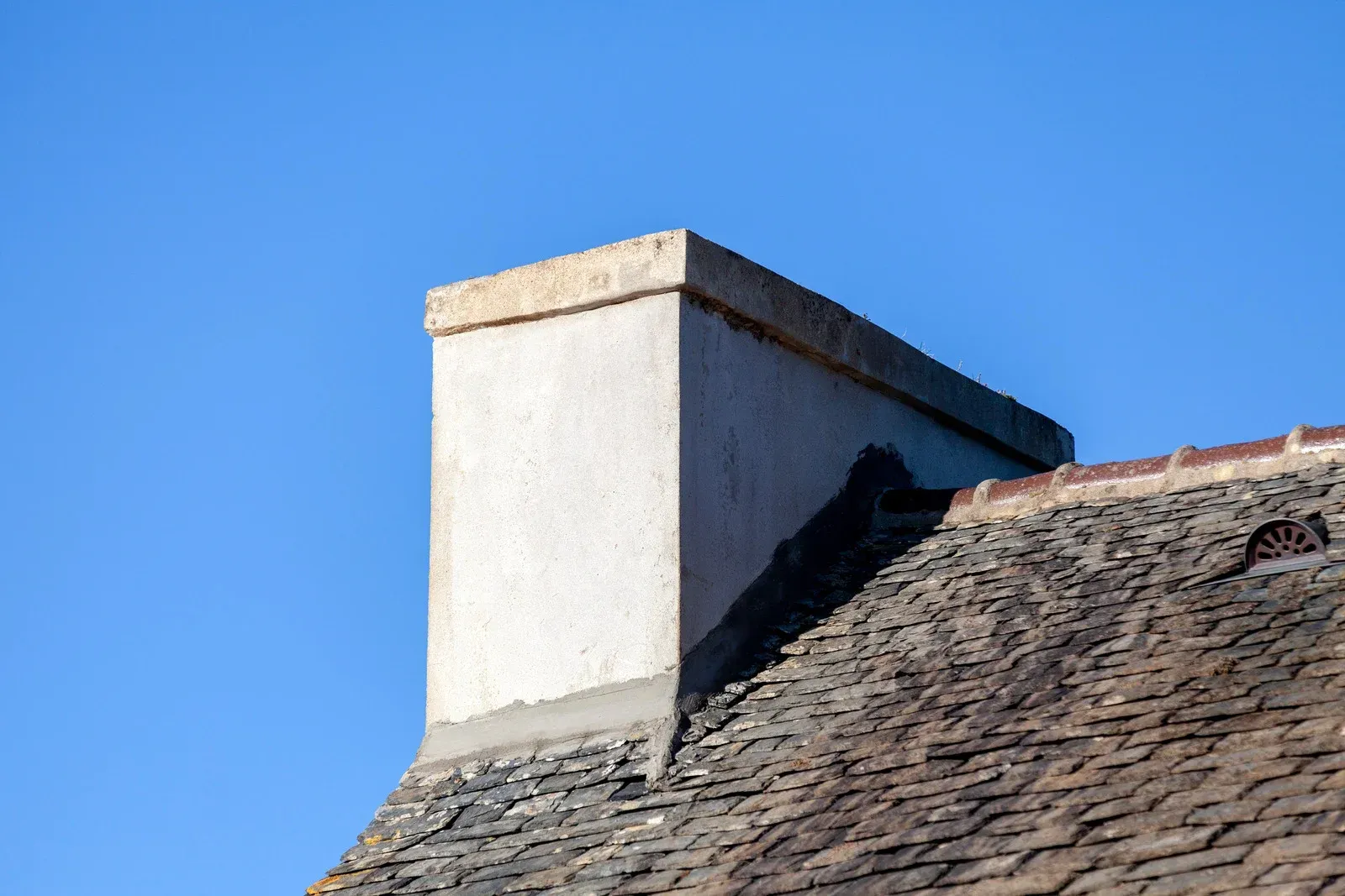 White chimney on a weathered, shingled roof against a clear blue sky.