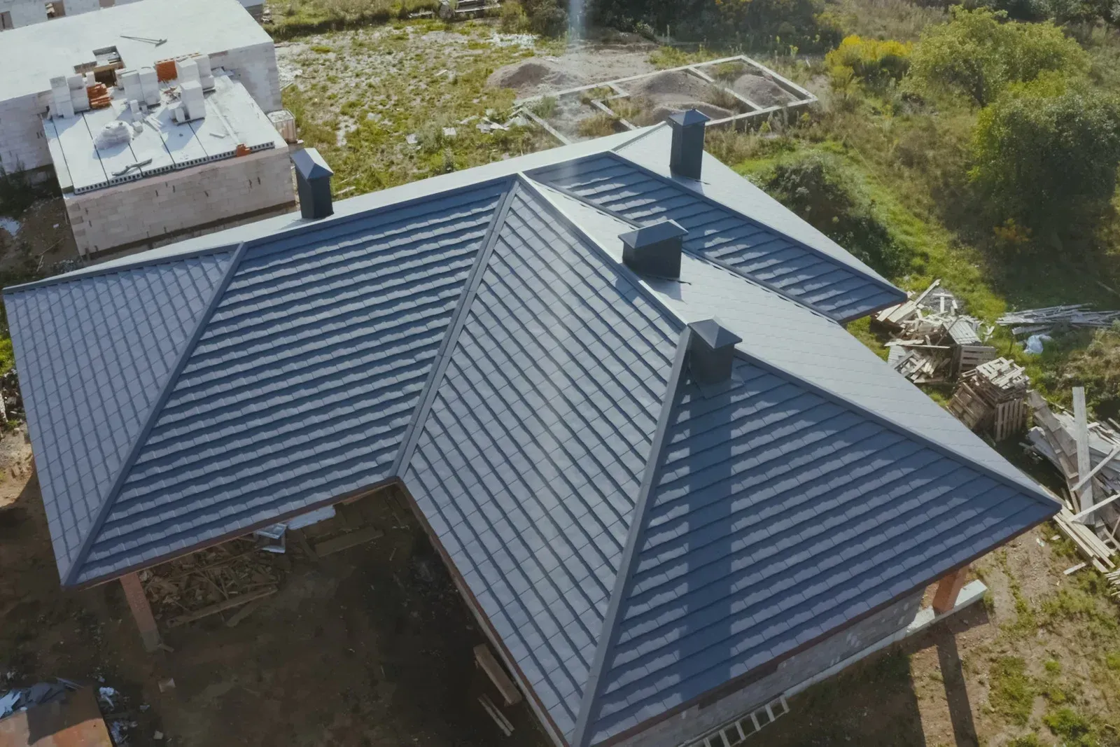 Drone shot of a house with a dark gray metal roof, three chimneys, and surrounding construction materials.