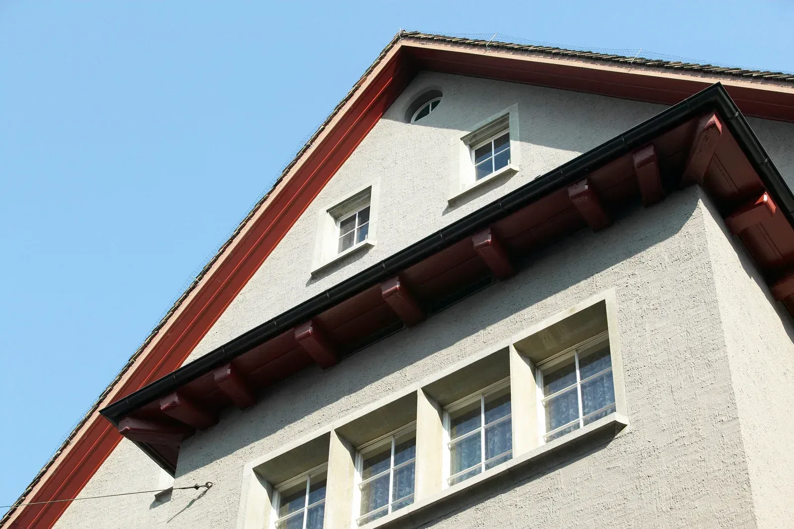 Close-up of a building's triangular roof with red trim, small windows, and light gray stucco walls against a blue sky.