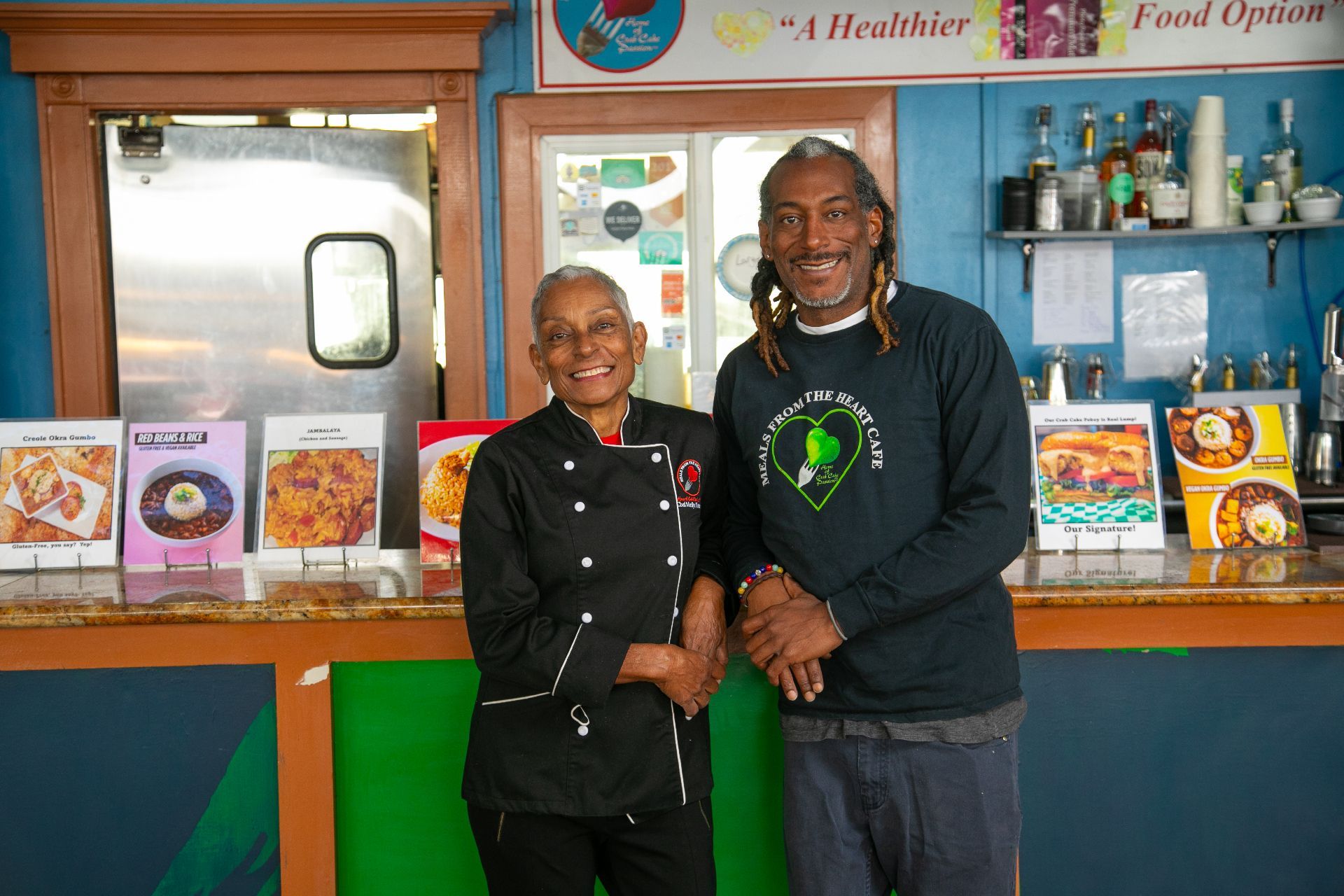 Two people stand smiling behind a counter in a health food shop, with jars, books, and packaged goods on display.