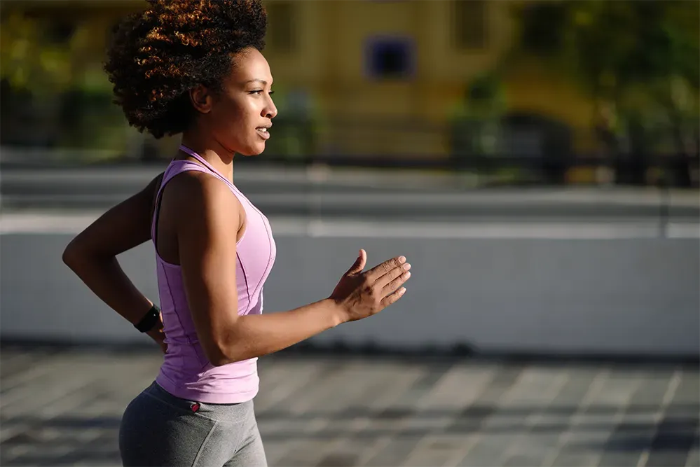 Woman jogging outdoors in a purple tank top and gray leggings, with blurred urban background