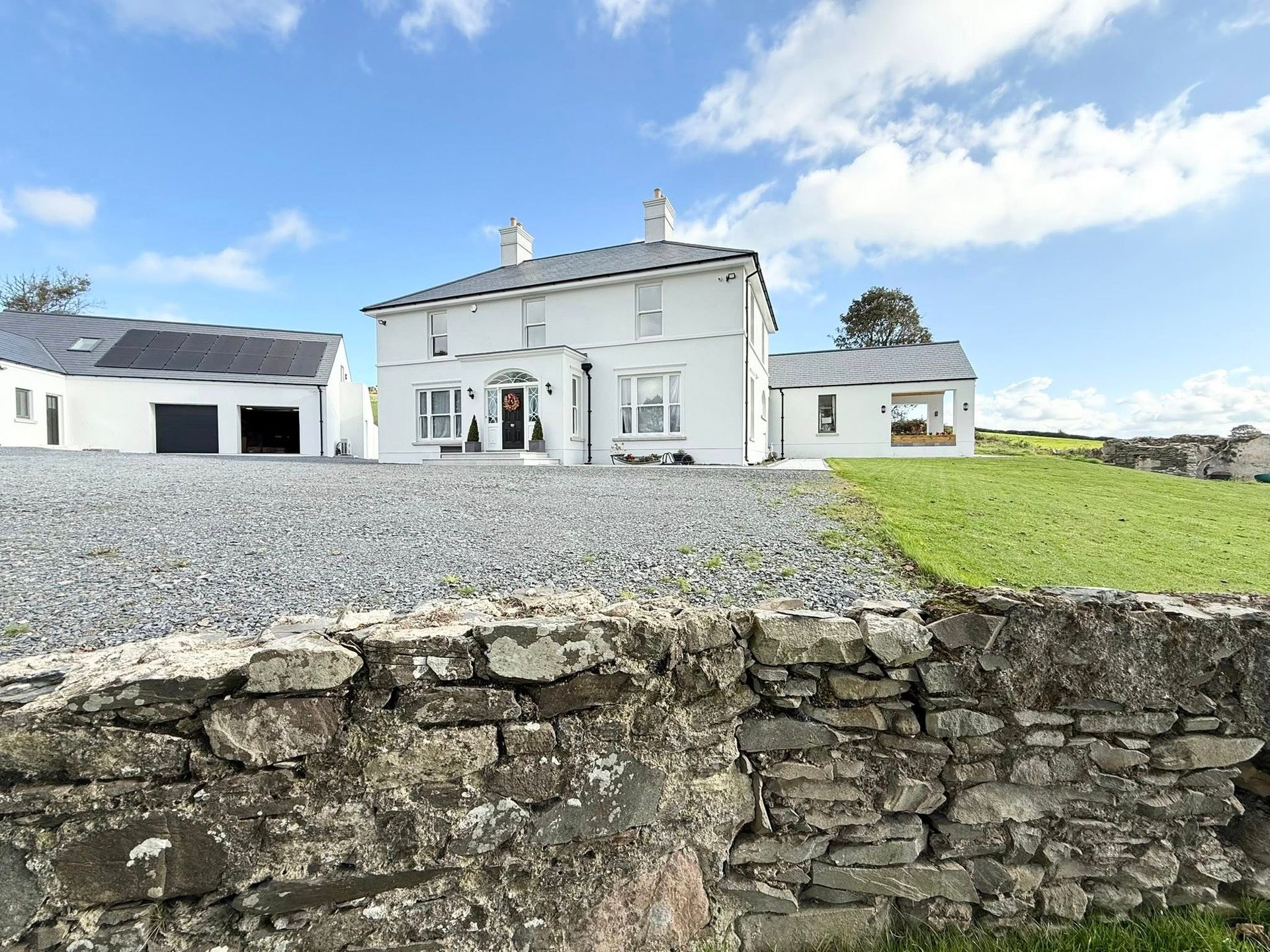 White house with outbuildings, gravel driveway, and stone wall under blue sky.