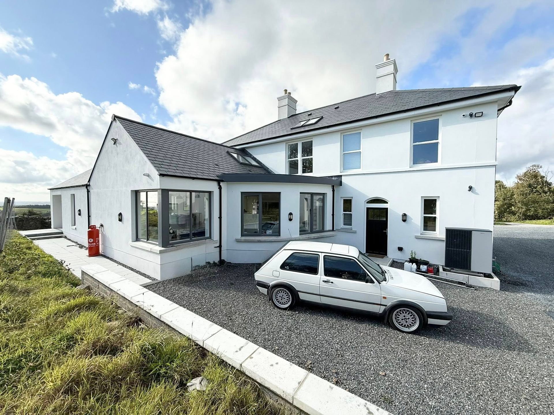 White house with a white car parked in front, on a gravel driveway, against a cloudy sky.