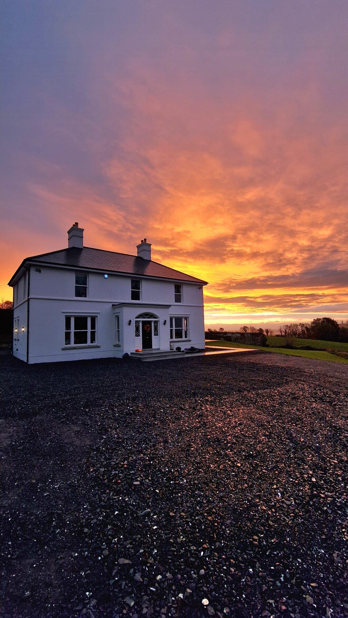 White two-story house with dark roof at sunset. Orange and purple sky over gravel driveway.