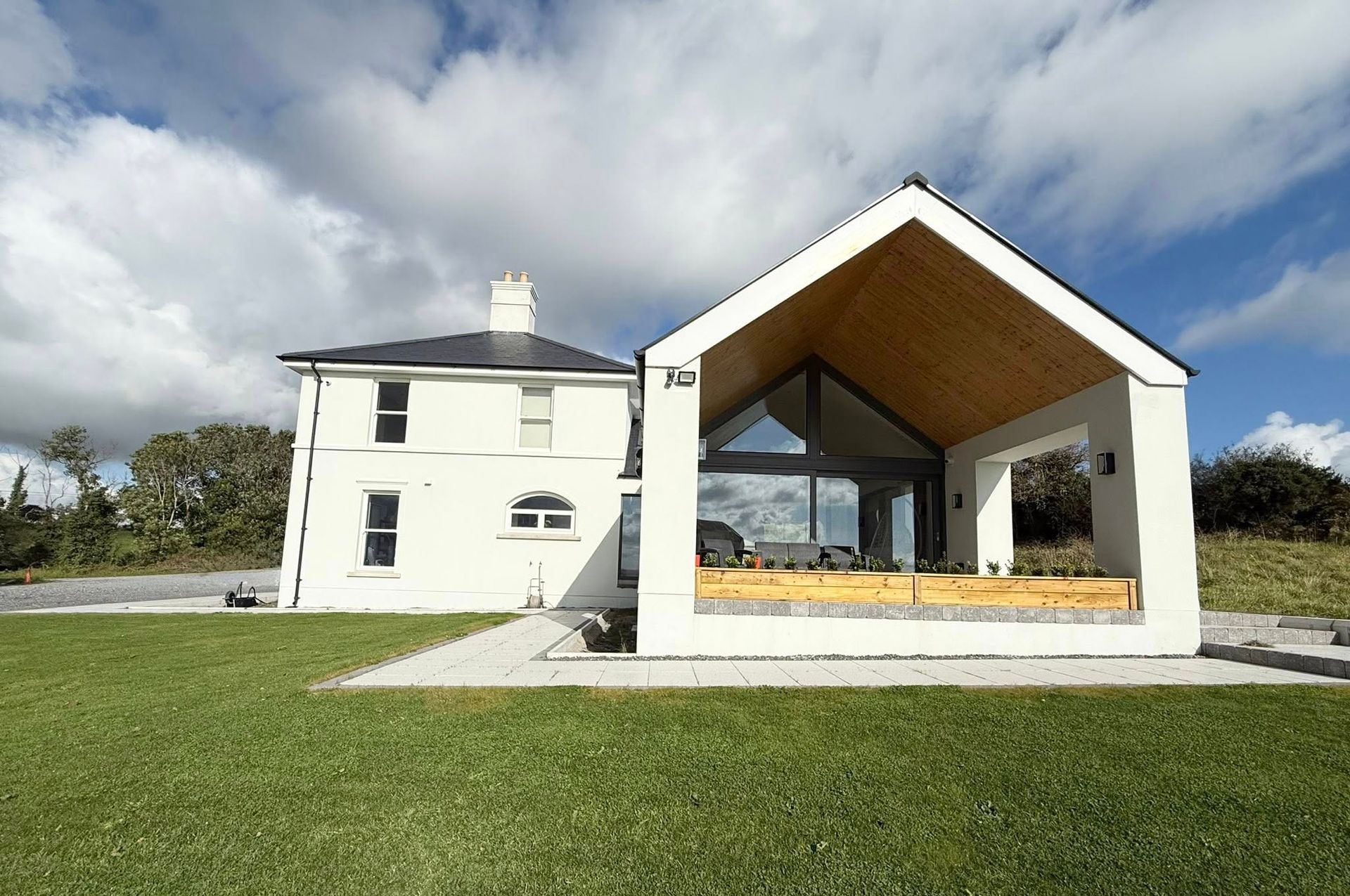 White house with modern glass extension, green lawn, blue sky with clouds.