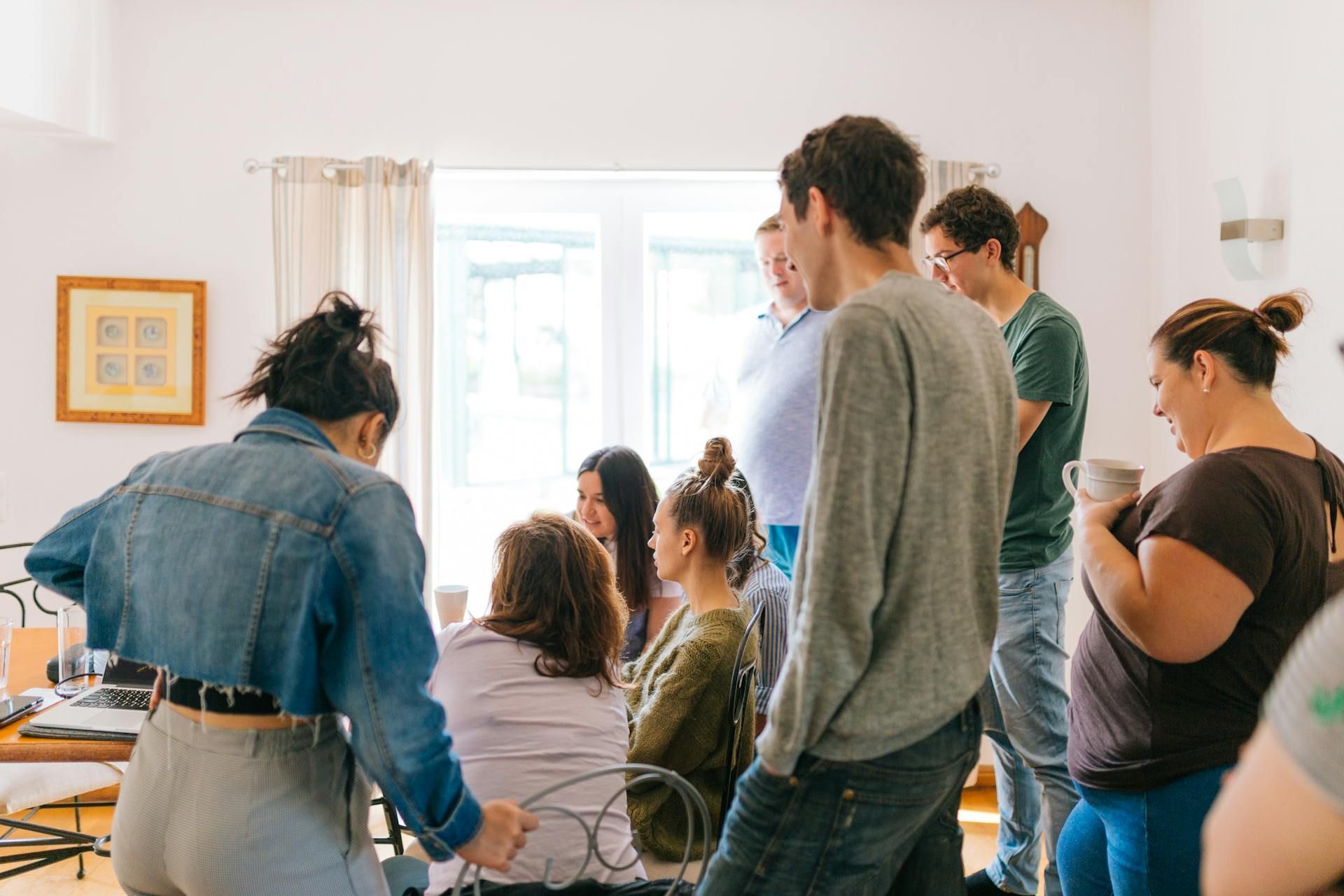 People gathered indoors, near a window, talking and drinking from mugs. Bright, natural light.