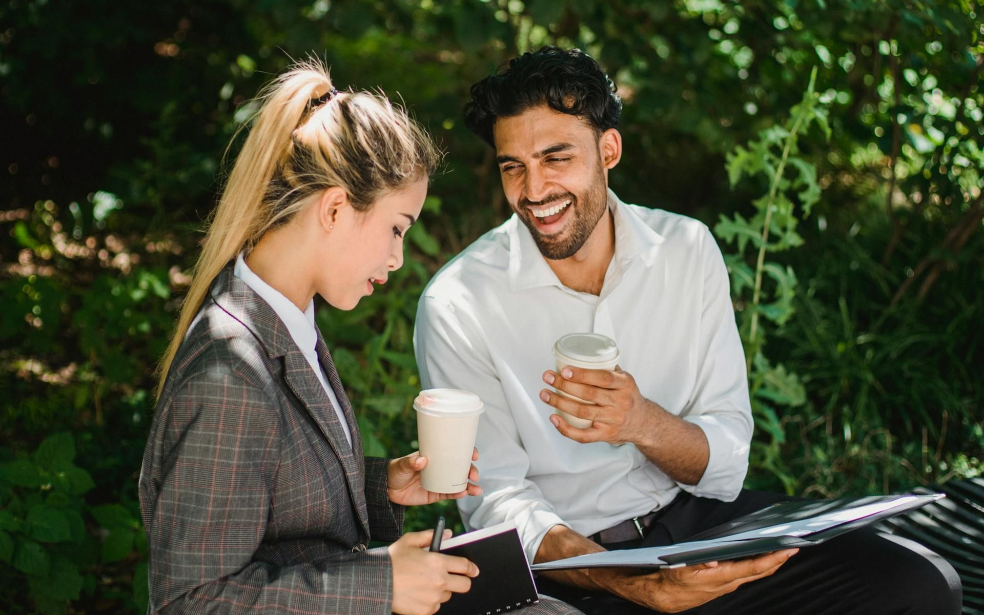 Two people sitting on a bench under trees, looking at a notepad and smiling. They are holding coffee cups.