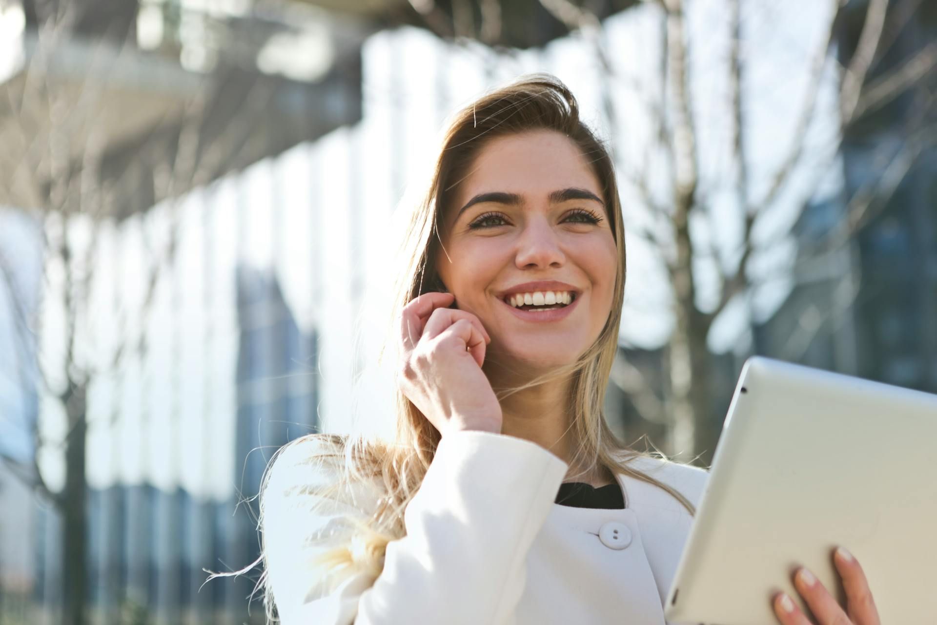 Woman in a white jacket, smiling, holding a tablet, and talking on the phone outside a building.