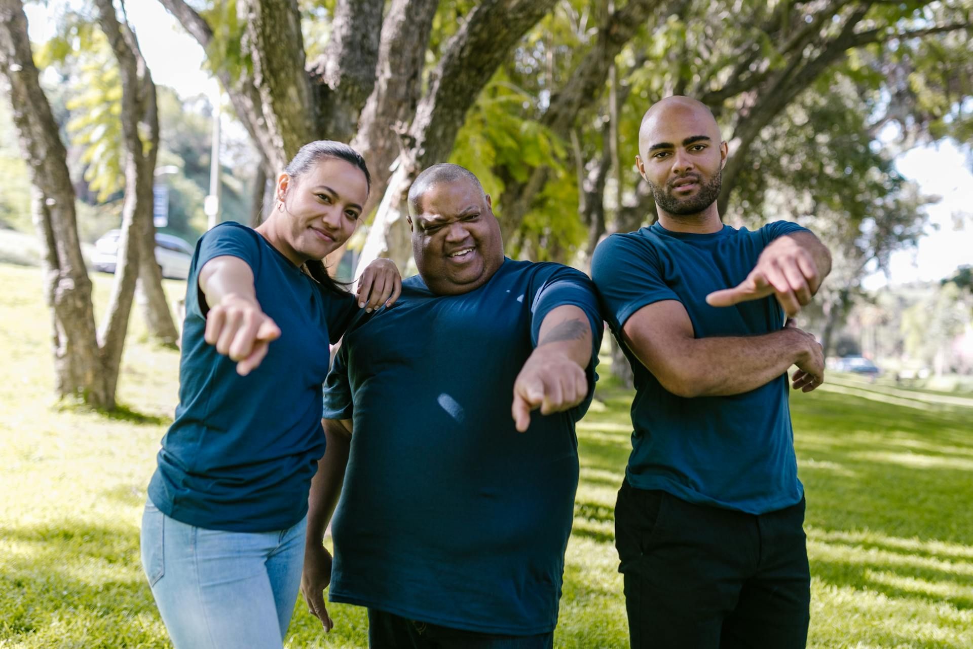 Three people in blue shirts pointing at the viewer, standing in a park setting.