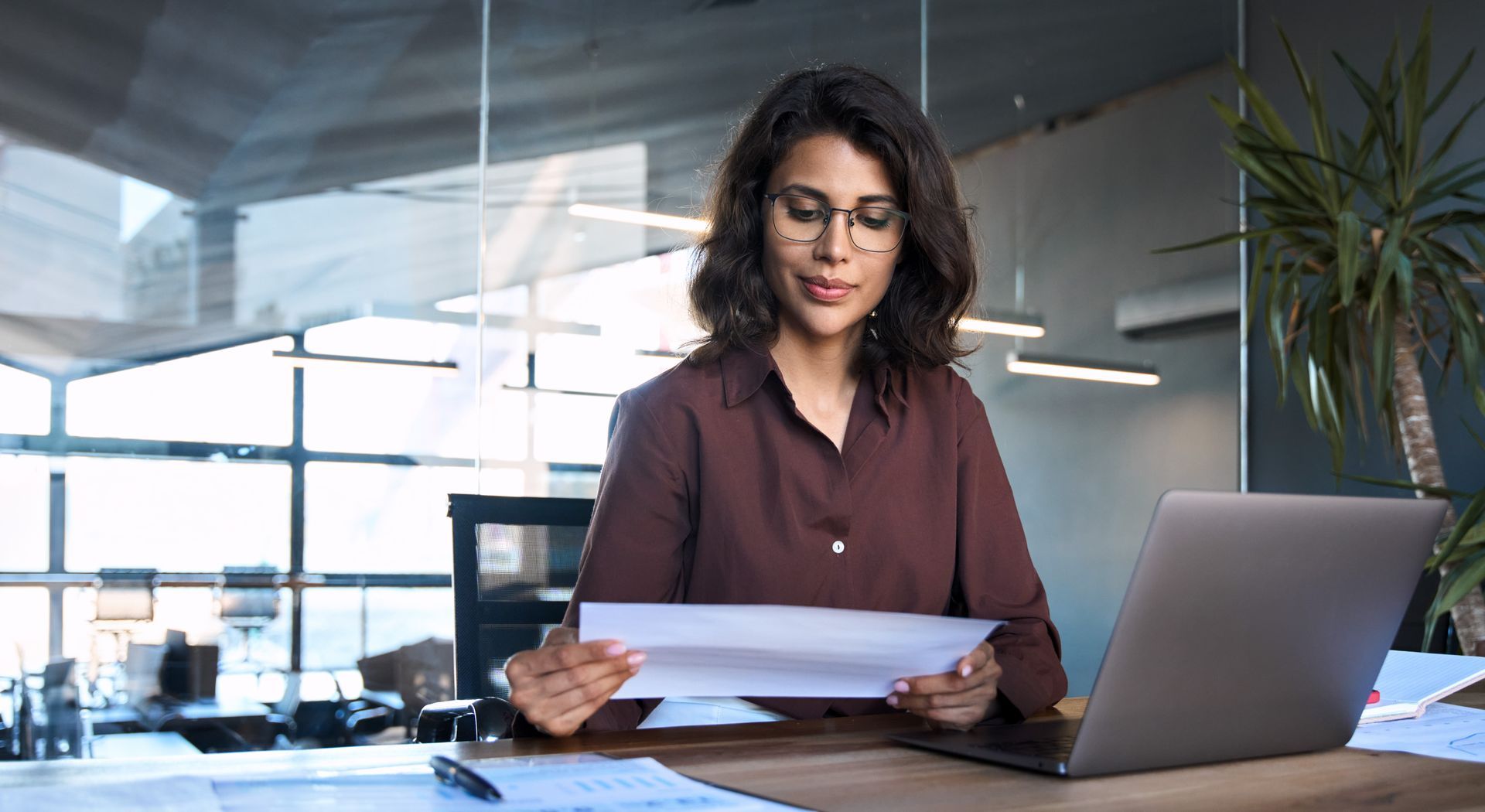 Woman with glasses in a brown shirt examines papers at a desk with a laptop in a modern office.