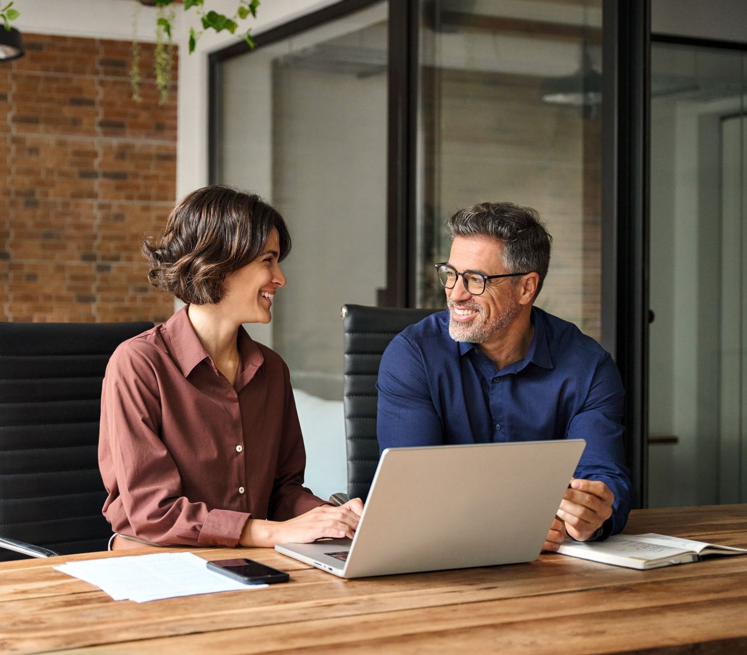 Woman and man smiling at each other while working at a laptop together in an office setting.