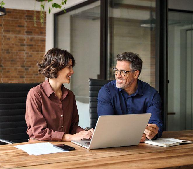 Woman and man smiling at each other while working at a laptop together in an office setting.