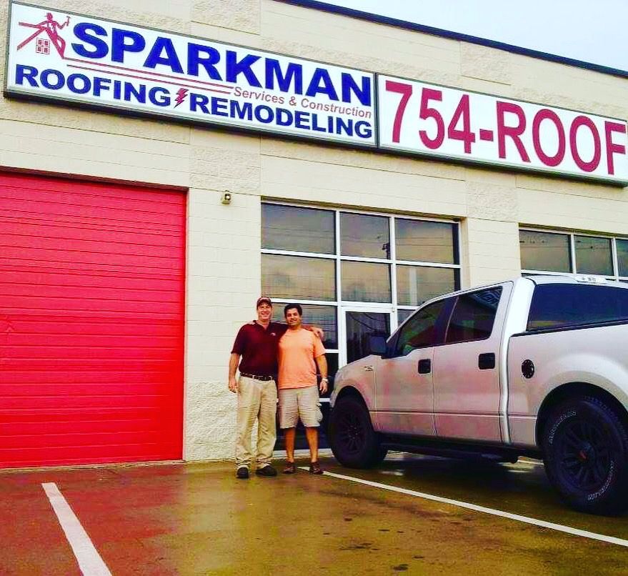 Two people stand in front of Sparkman Roofing and Remodeling, next to a silver truck in a parking lot.