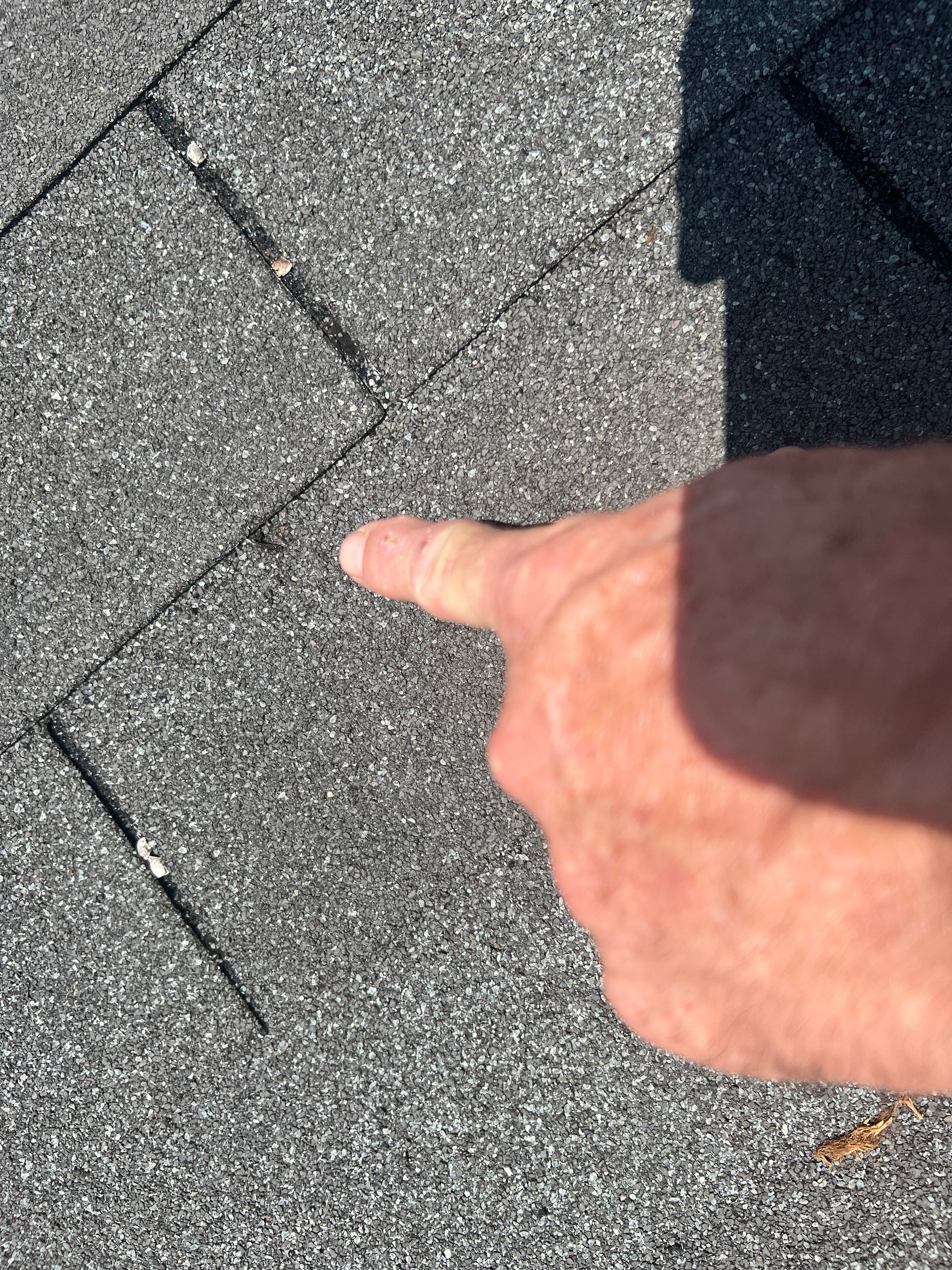 A finger points to a small crack in the textured, gray surface of an asphalt roof shingle.