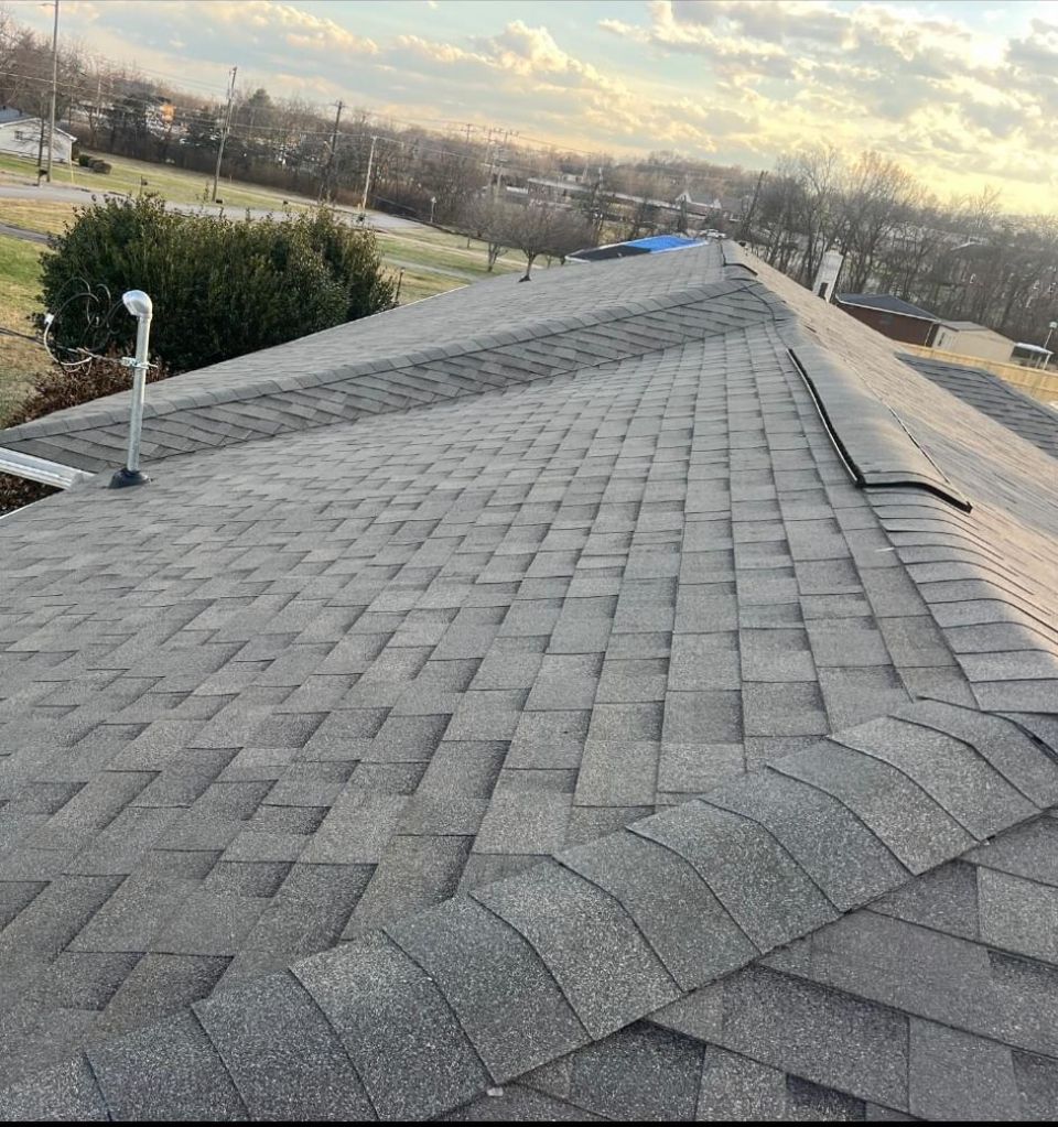 A view looking down the ridge of a gray asphalt shingle roof on a sunny day with a suburban landscape in the background.