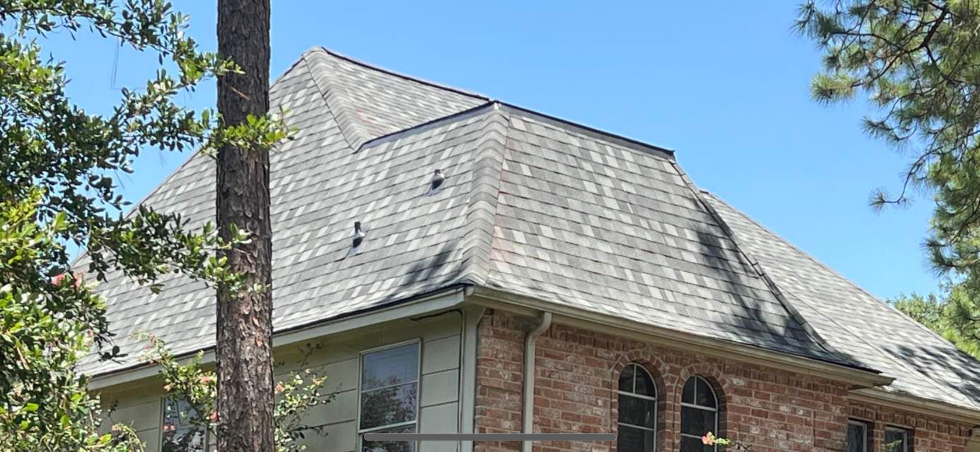 A tan brick house with a complex grey shingled roof under a clear blue sky, partially obscured by green trees.
