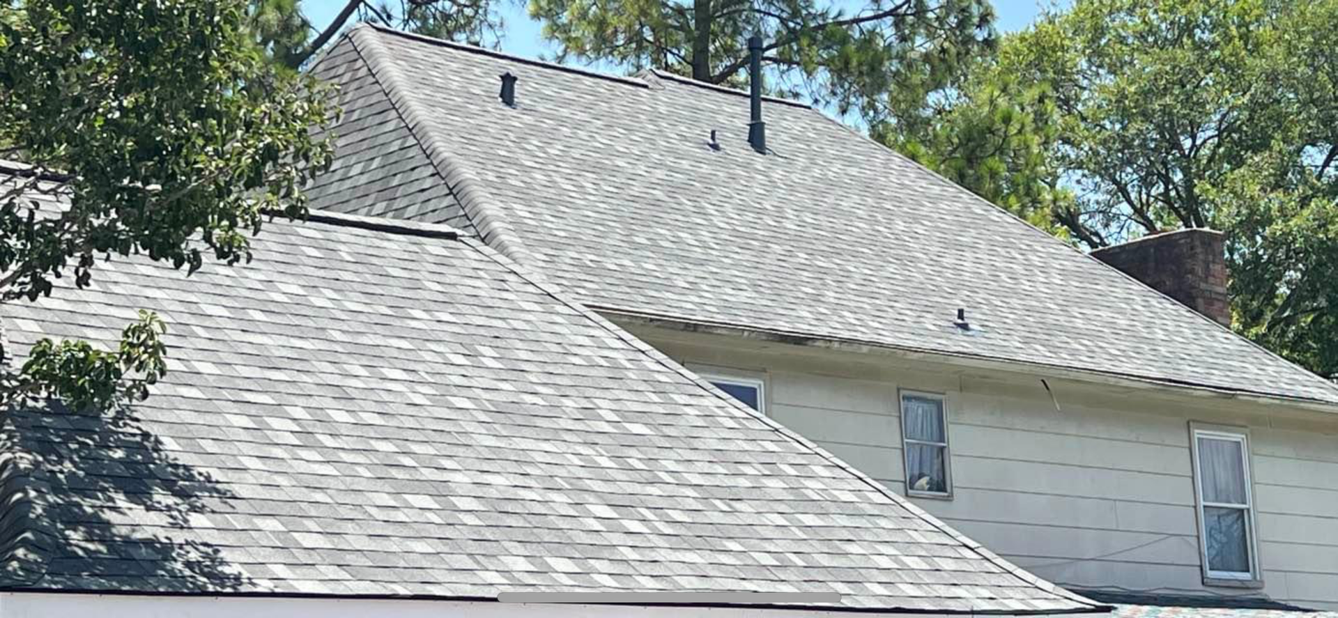 A side view of a house roof with grey asphalt shingles, featuring two roof planes and white siding with two windows.
