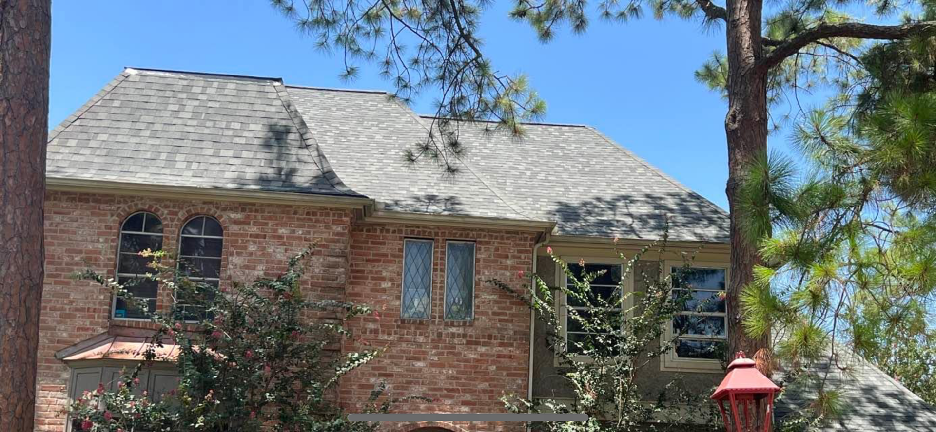 Two-story brick house with a gray shingled roof, arched windows, and pine trees under a clear blue sky.