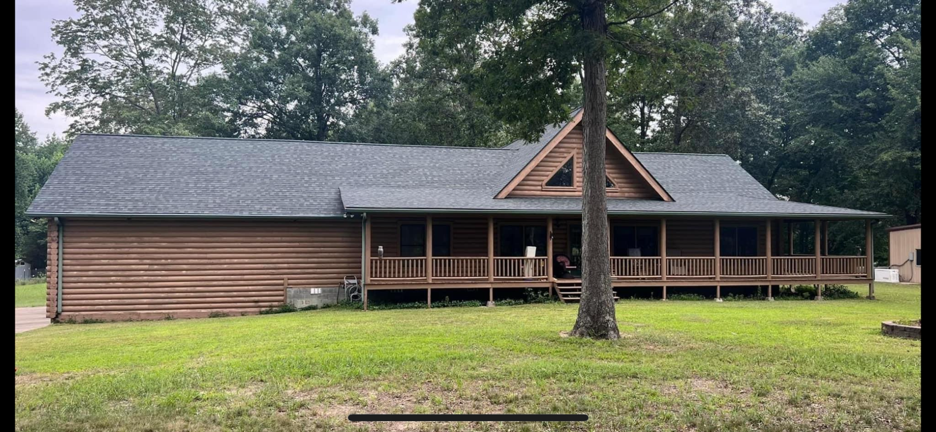 A sprawling, single-story log cabin with a wide covered front porch, grey shingled roof, and lush green lawn in the woods.