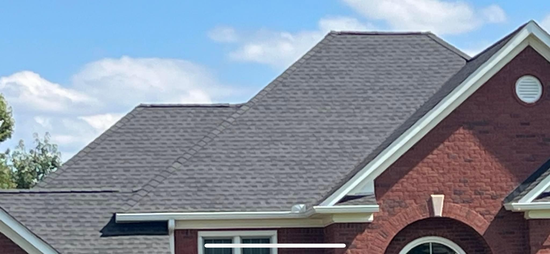 The dark gray shingled roof of a brick house under a blue sky with light clouds.