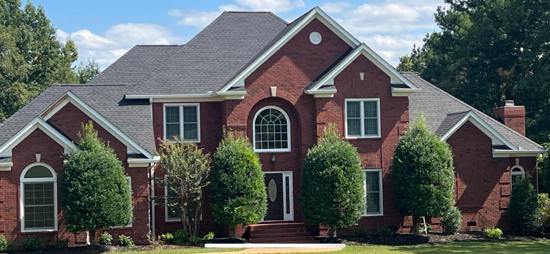 A two-story red brick house with a gray shingled roof, multiple gables, and several manicured shrubs in the front yard.