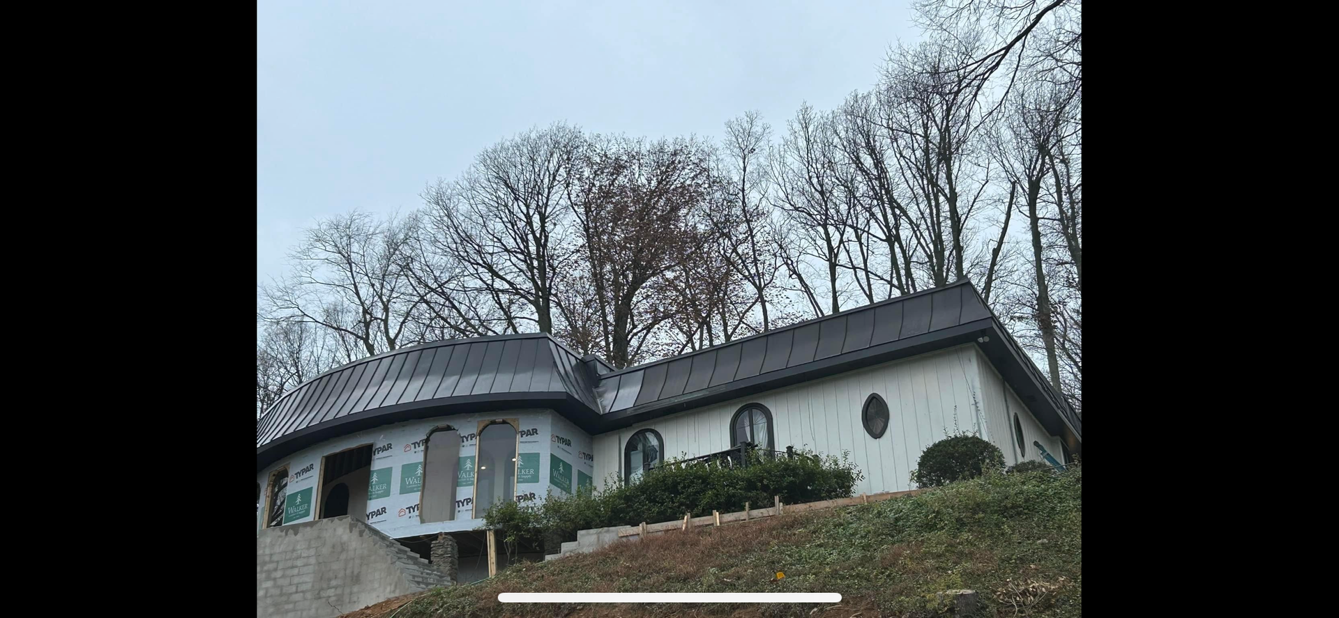 A white house with a dark metal roof sits on a hill against a backdrop of bare trees under an overcast sky.