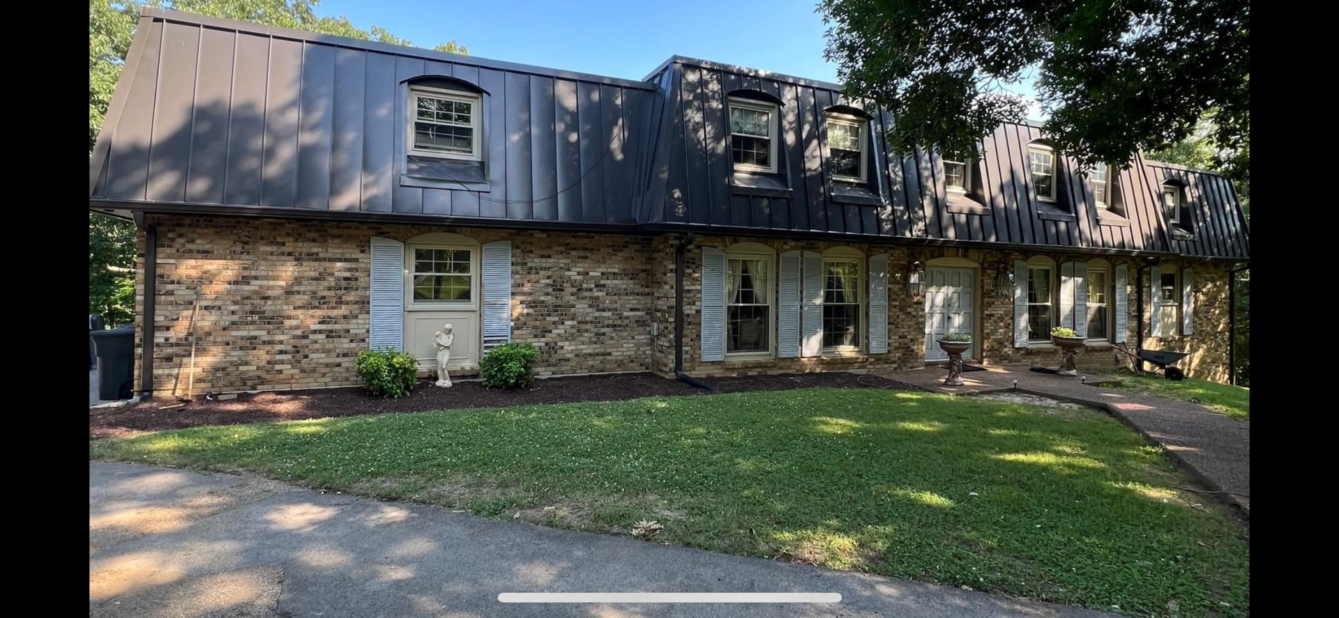 A two-story stone house with a dark metal mansard roof, dormer windows, and a front lawn under a bright, sunny sky.