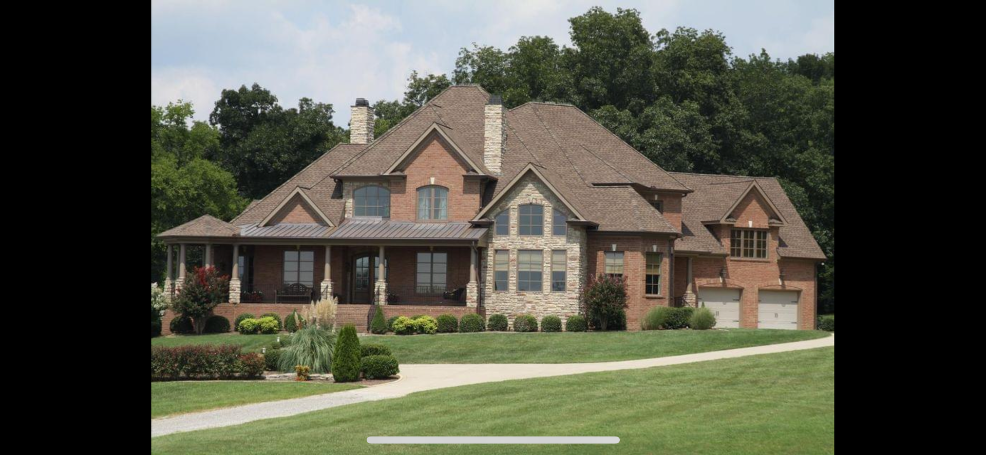 A large brick house with stone accents, a covered porch, and a gravel driveway set against a backdrop of green trees.