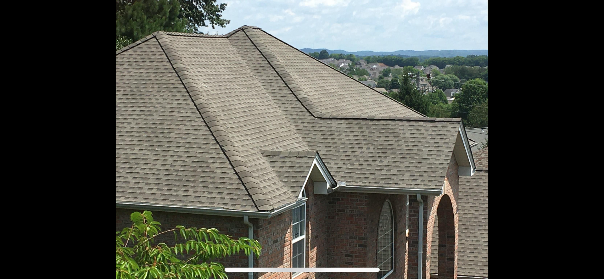 A residential brick home with a multi-gabled, brown shingled roof under a partly cloudy sky.