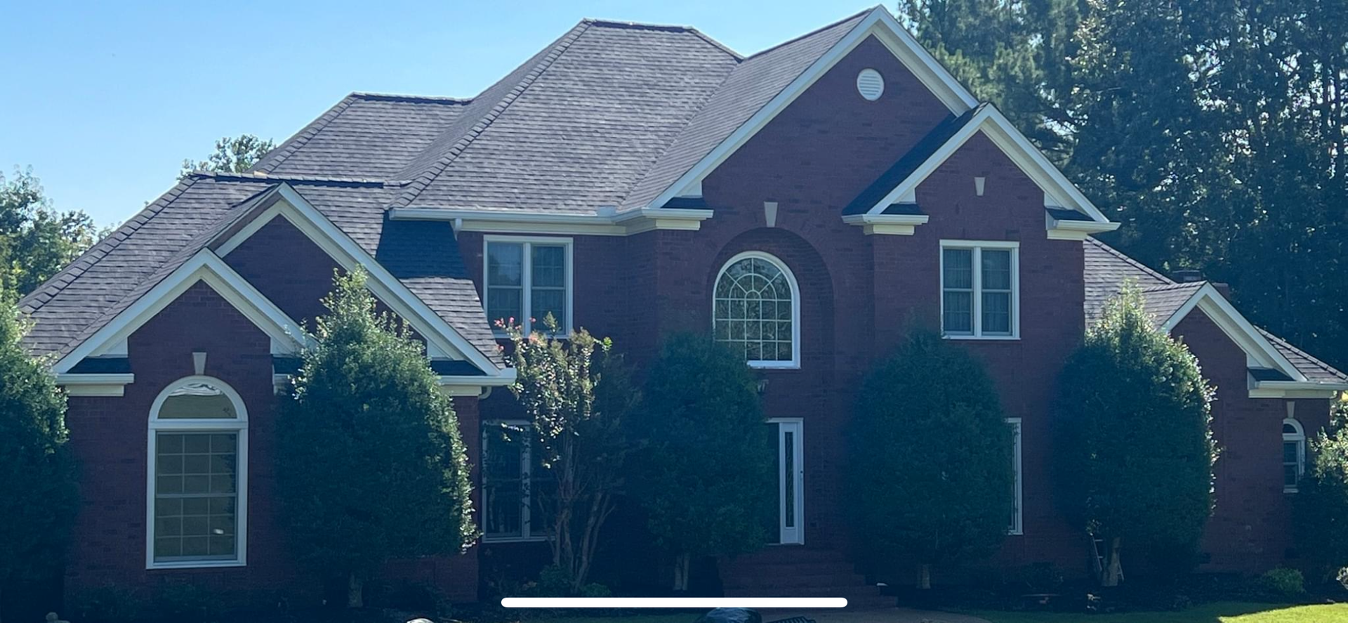 A two-story red brick house with a dark shingled roof, arched windows, and surrounding green trees under a blue sky.