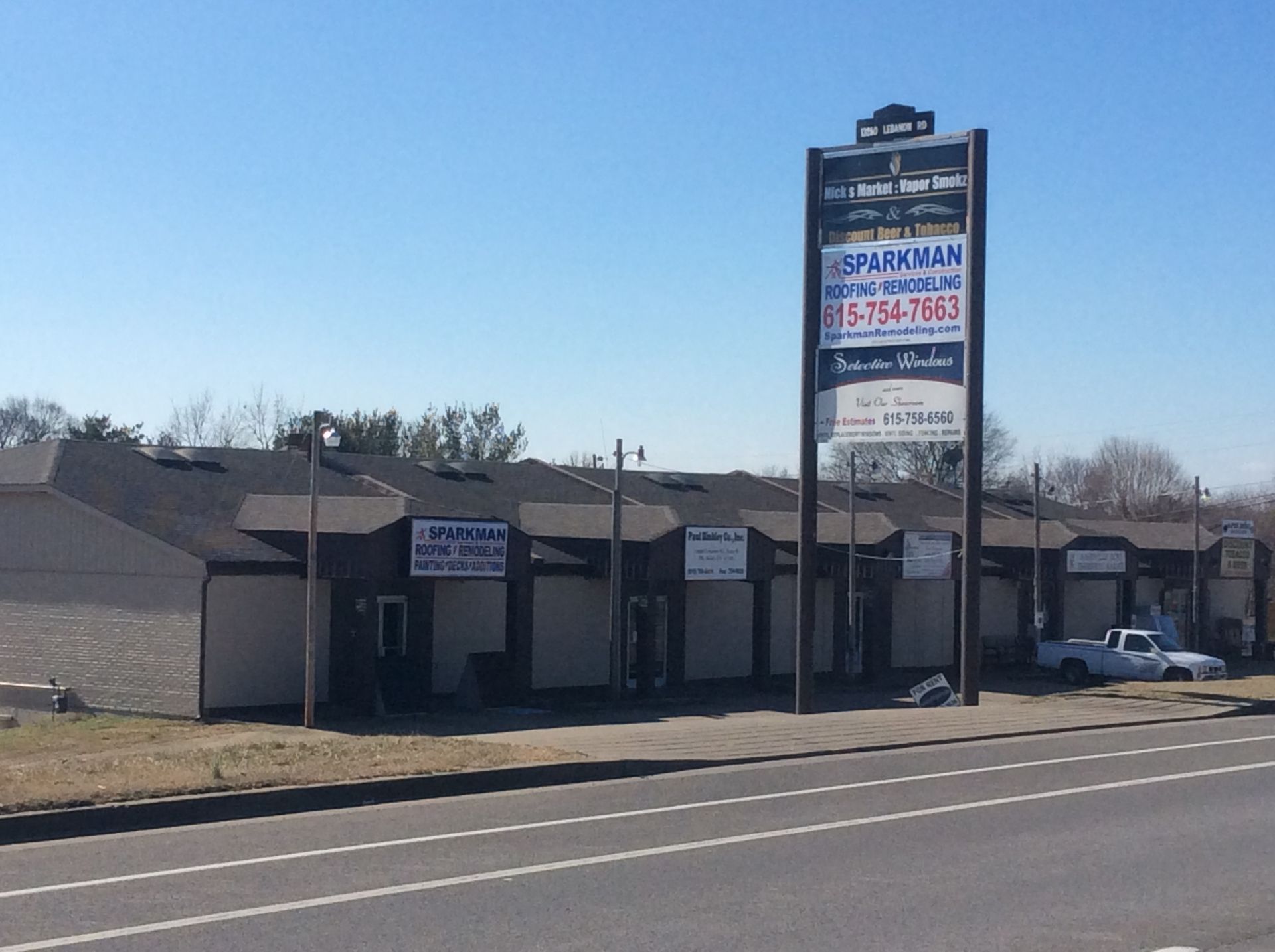 A tan commercial strip mall with a prominent freestanding signage pole by a road under a clear blue sky.
