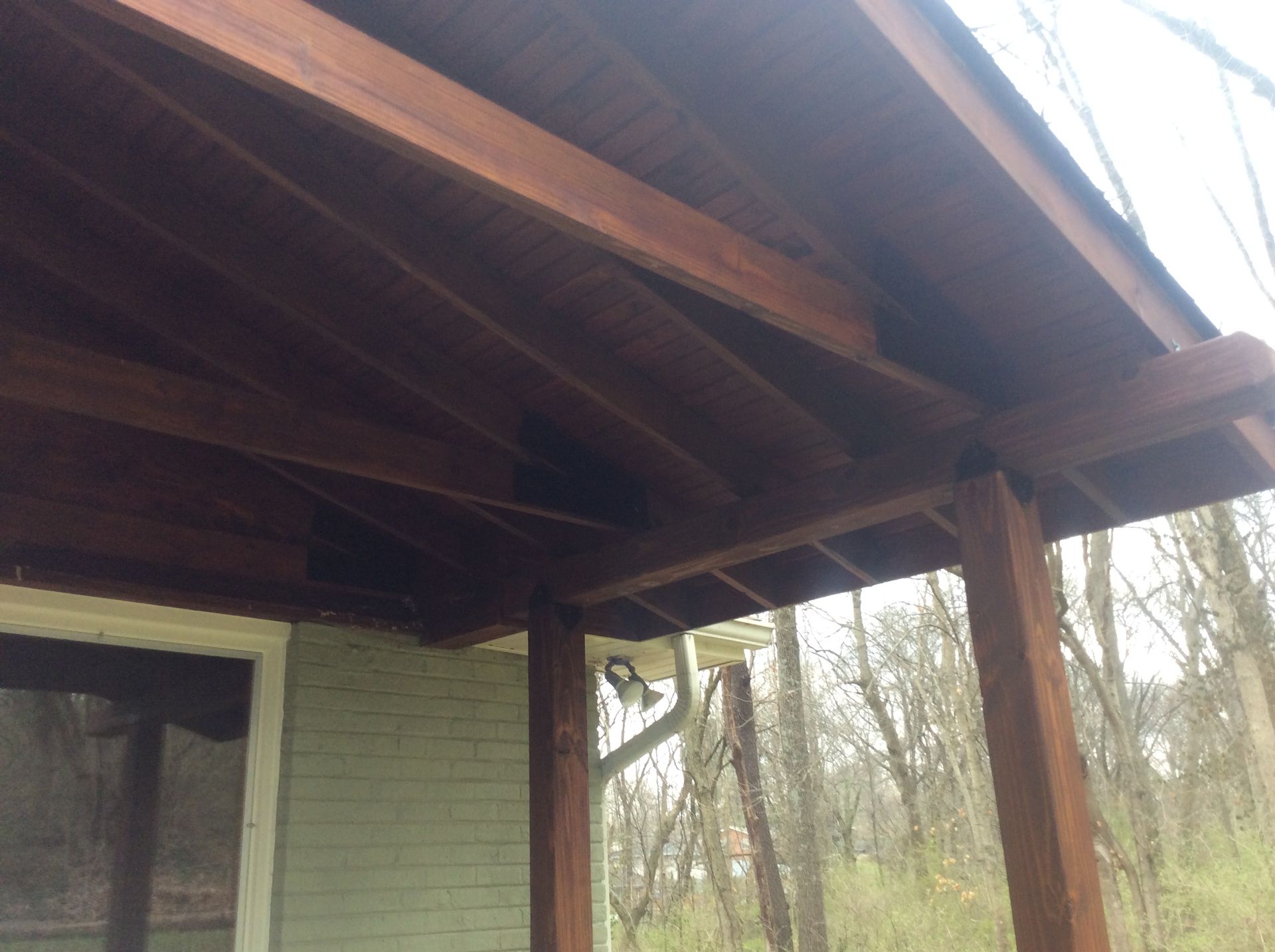 A low-angle view of a wooden porch ceiling and roof structure attached to the side of a light-colored brick house.