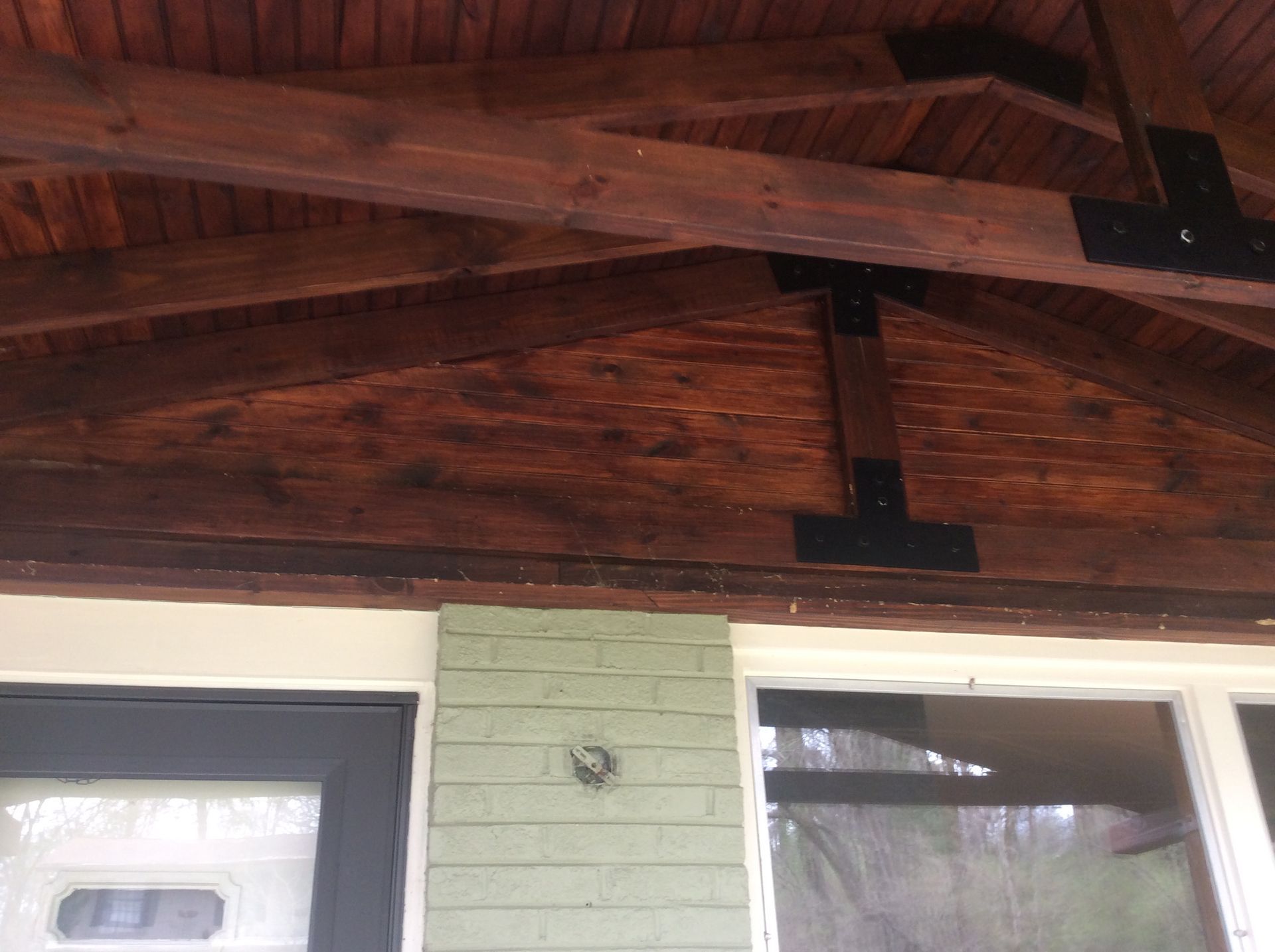 A wooden porch ceiling with dark stained beams and black metal brackets above a front door and green brick wall.