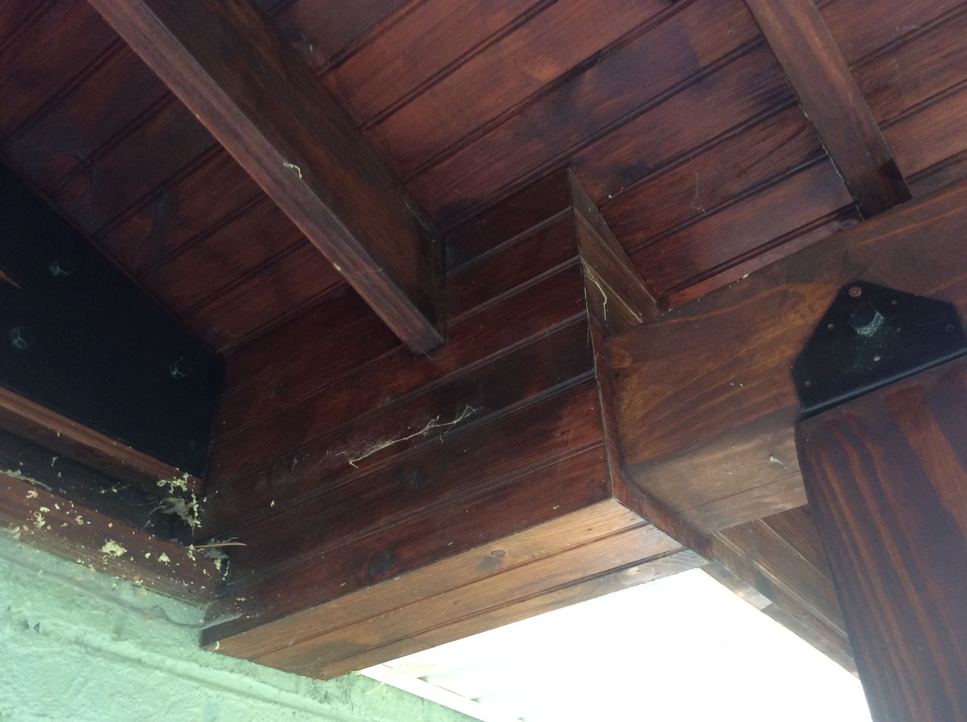 Close-up view of wooden deck framing, showing beams, rafters, and a metal bracket connection against a textured wall.