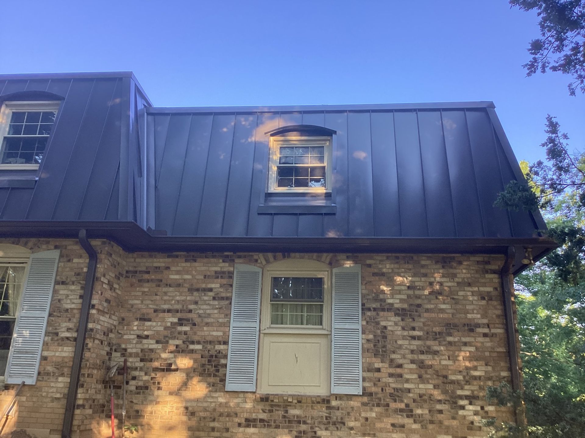 A two-story brick house with a dark metal mansard roof and two dormer windows under a clear blue sky.