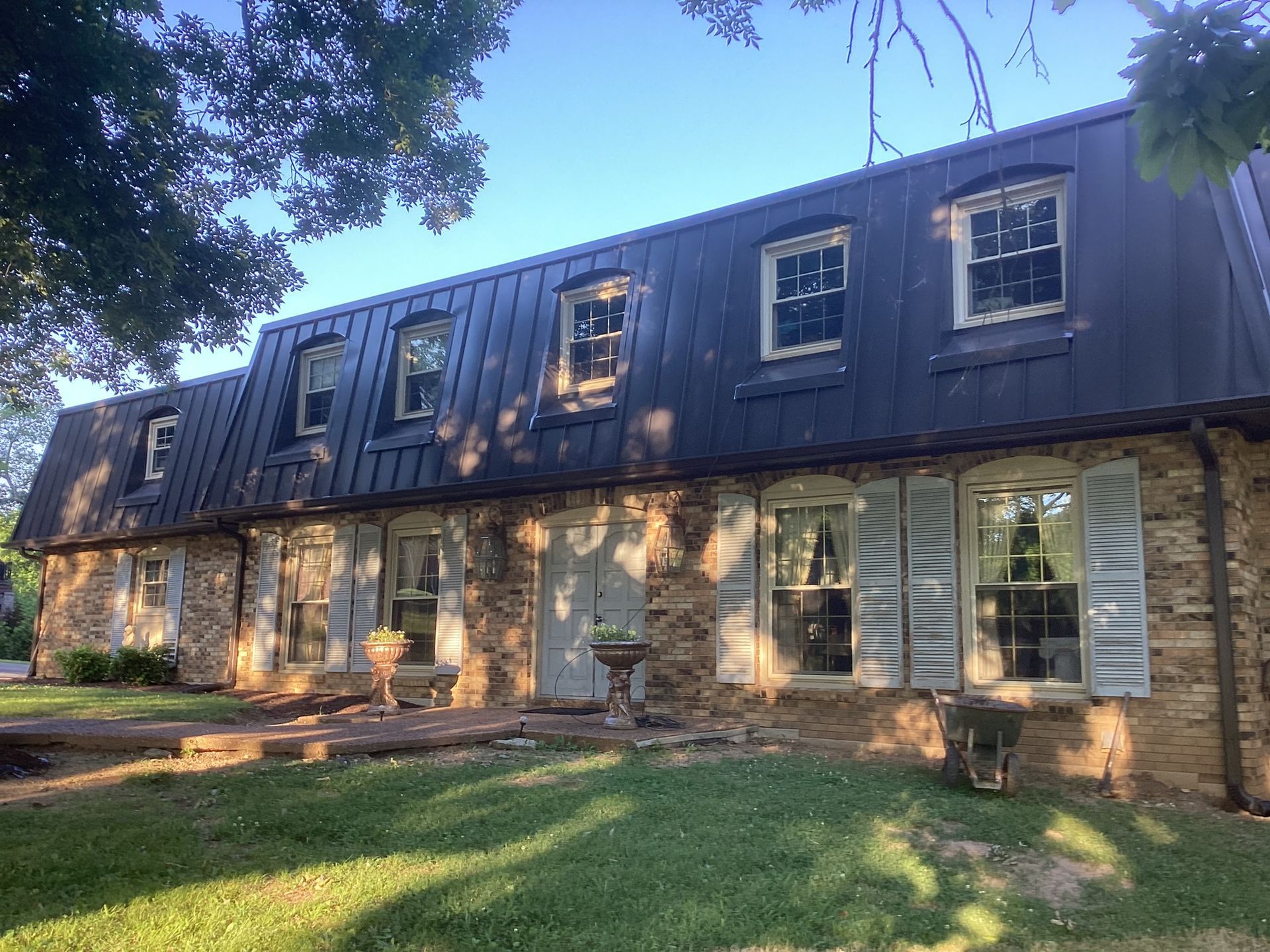 Two-story stone house with a mansard roof, dormer windows, and a front lawn on a sunny day.