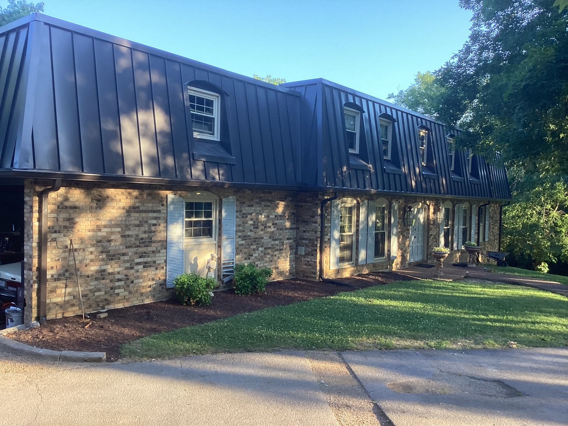 A long, two-story house with a stone brick facade and a dark metal mansard roof, surrounded by a lawn and trees.