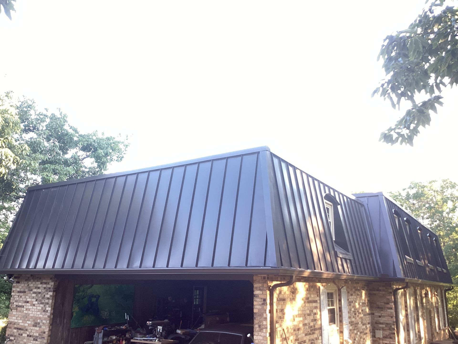 A stone structure with a dark, standing-seam metal roof set against trees under a bright sky.