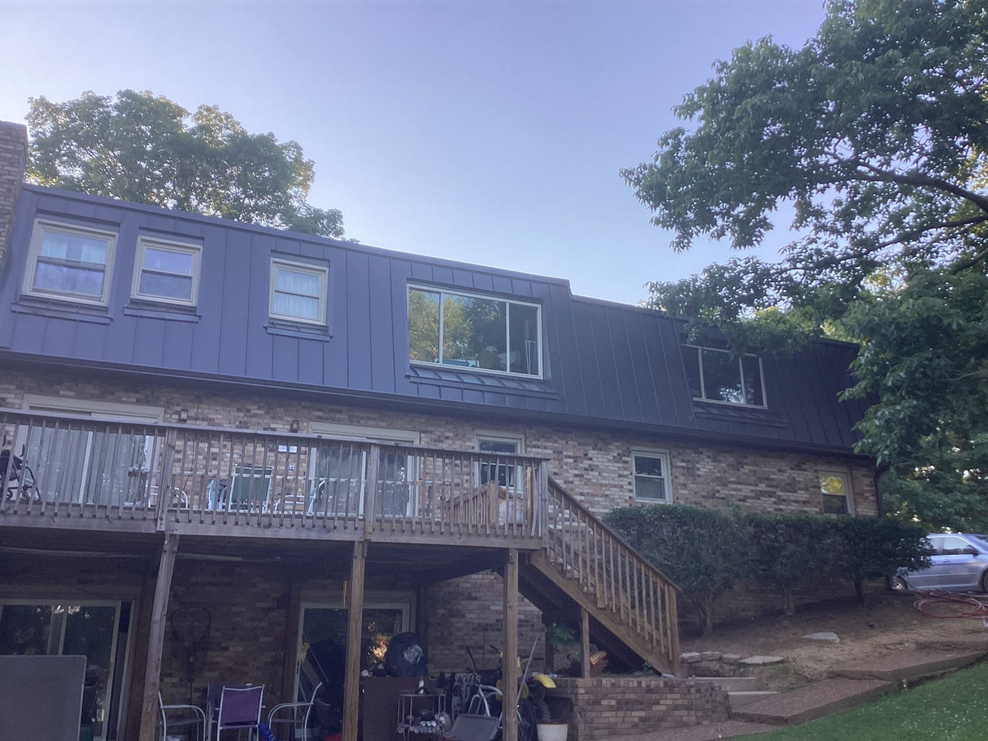 A stone two-story house with a dark metal mansard-style roof, a wooden deck, and a staircase, surrounded by trees.
