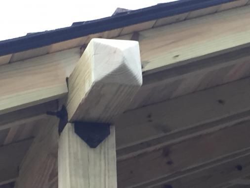 A wooden post with a pyramid-shaped top cap, connected by a black metal bracket to a horizontal beam under a roof edge.