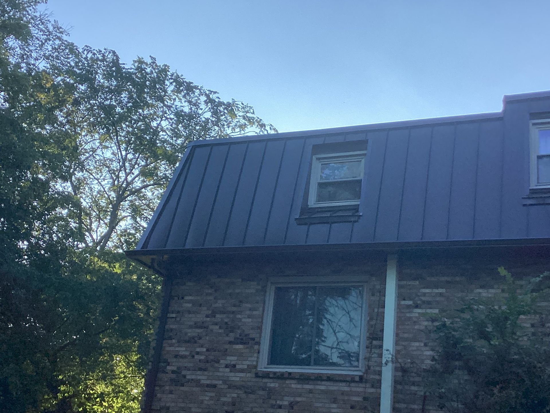 A dark metal roof with a dormer window on a brick house, partially obscured by green trees under a clear blue sky.