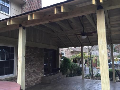 A wooden patio cover attached to a brick house, featuring a ceiling fan and support posts over a concrete patio.