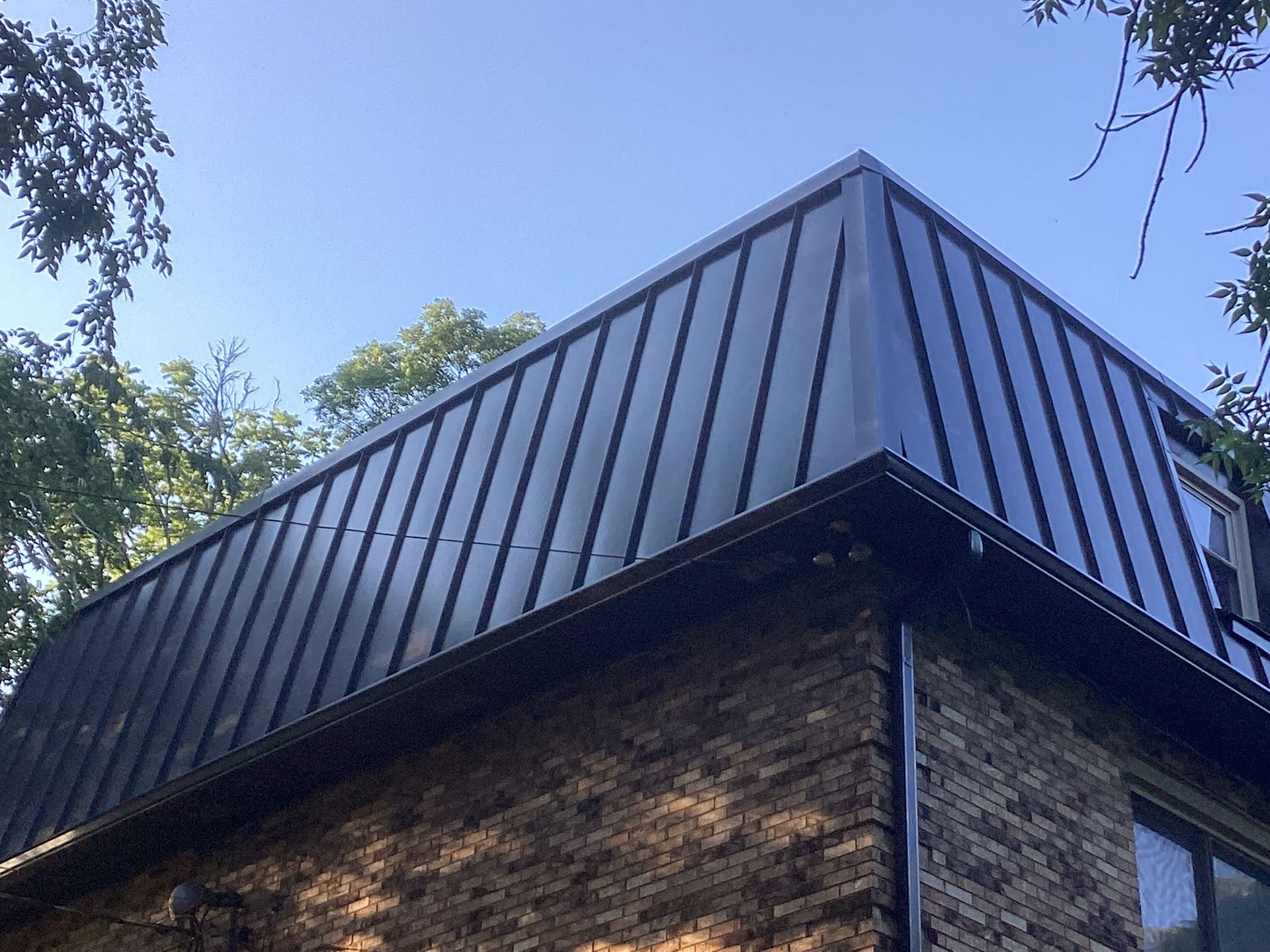 A dark, vertical-seam metal roof sits atop a brick building against a bright blue sky.